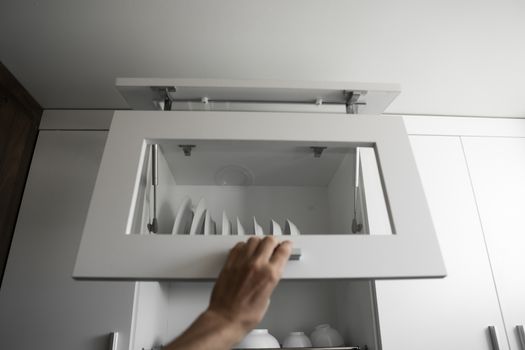 Hand holds the door of the dryer with utensils. Dish drying metal rack with white clean plates. Traditional comfortable kitchen. Open white dish draining closet with wet dishes of plates, bowls