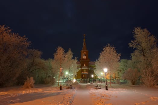 The building of the Orthodox Church at night surrounded by trees in the snow illuminated by lanterns.