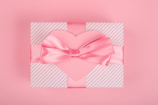 Valentine Day gift in a box wrapped in striped paper and tied with silk ribbon bow and heart shapes greeting card on pink background with copy space for text