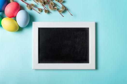 Orthodox Easter concept. Colorful eggs and pussy willow branches on blue background with empty chalkboard. Copy space for greetings, text or design. Top down view or flat lay