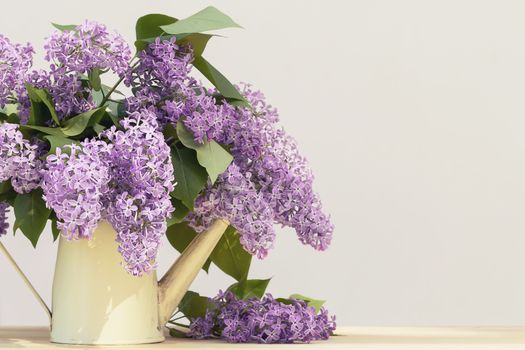 Vase in the form of an old vintage watering can with a bouquet of lilacs on a wooden table on a light background.