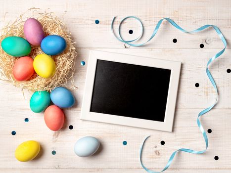 Easter concept. Colorful eggs on white wooden background with empty chalkboard. Copy space for greetings, text or design. Top down view or flat lay