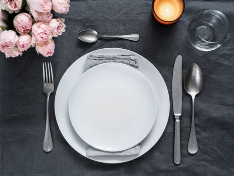 Beautiful table setting on gray linen tablecloth. Festive table setting for wedding dinner with pink spray roses and scented cabdle. Holiday dinner with white plates. Dinner, utensil. Copy space