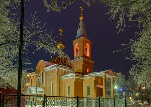 The building of the Orthodox Church at night surrounded by trees in the snow illuminated by lanterns.