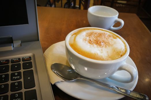 Cup of Cappuccino and laptop on wooden table, Cafe. Laptop pc computer open keyboard next to cappuccino coffee cup on light brown wooden table in cafe wit nobody