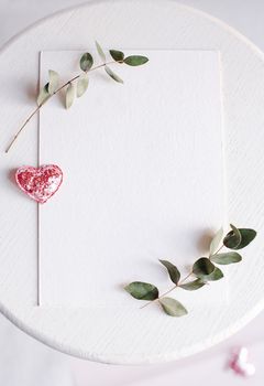 Background with copy space blank on table with glitter heart, eucalyptus branch, flowers and leafs. White paper top view, flat lay, minimal style. Moke up card