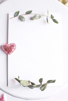 Background with copy space blank on table with glitter heart, eucalyptus branch, flowers and leafs. White paper top view, flat lay, minimal style. Moke up card