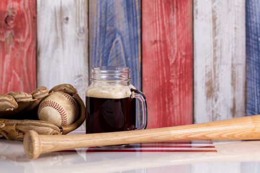 Front view of a glass of cold dark beer with a worn baseball mitt, bat and ball.  Faded wooden boards painted red, white and blue in background with USA flag underneath.  