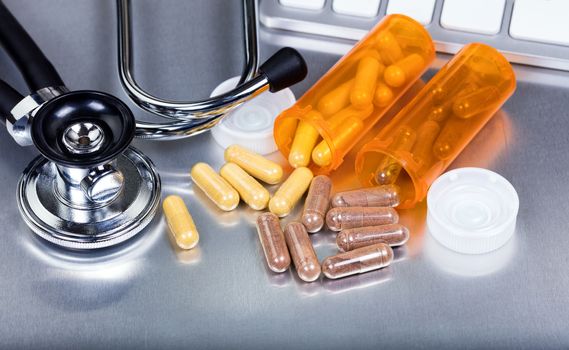 Close up of capsules spilling out of containers with stethoscope and keyboard in background on stainless steel surface. 