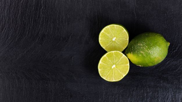 Overhead view of fresh limes, whole and sliced, on black slate with available copy space on left side