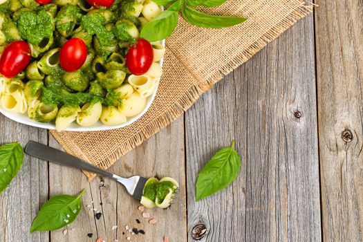 Top view of basil pesto with shell shaped pasta and cherry tomatoes on rustic wood. 