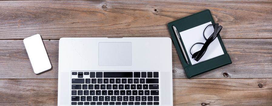 Flat view of working desktop consisting of laptop computer, smartphone, paper, pen and reading glasses