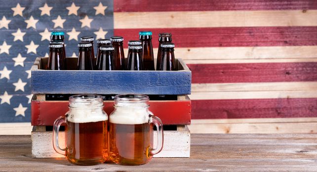 Two pint jars filled with beer, crate with unopen bottle and vintage wooden USA flag in background. Holiday party concept. 