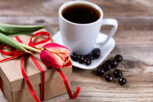 Single tulip resting on gift box with a cup of coffee and dark chocolate in background on rustic wood. Selective focus on front part of flower. 