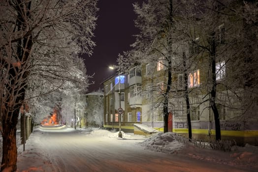 Night snowy street with trees in the light of lanterns and windows of houses painted with different colors.