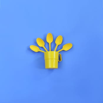 A yellow plastic mug and spoons lay in it on a bright blue background. The concept of a holiday, a picnic