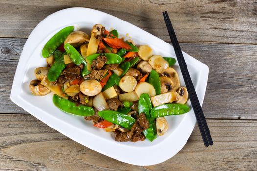 High angled view of Asian dish consisting of sliced juicy beef rice, onion, mushroom, green peas, and red pepper. Chopsticks on side of plate with rustic wood underneath. 