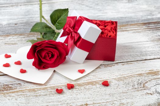 Open Valentines gift box filled with candies and single red rose on rustic white wood in close up view 
