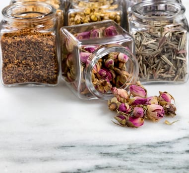 Front view of herbs and spices in glass jars with one jar spilling onto white marble stone. Selective focus on spilled rose buds. 