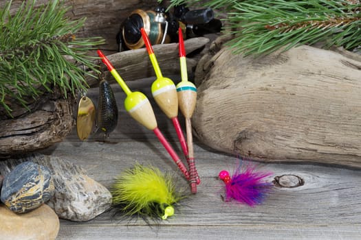 Horizontal image of fishing equipment consisting of floats, reel, lures, and feathered flies resting against aged driftwood, evergreen tree branches and rocks 