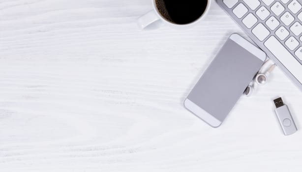 Overhead view of white desktop with partial computer keyboard, cell phone, coffee, thumb drive, and ear phone. 