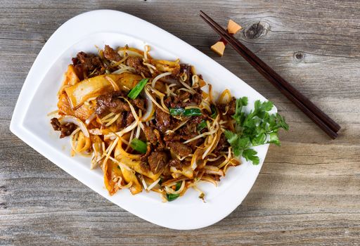 Top view of freshly cooked spicy beef, bean sprouts, onion, tofu with parsley as garnish and chopsticks in holder on rustic wood. 