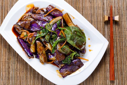 High angled view of cooked eggplant, onion and basil leaves in juicy sauce. Chopsticks in holder with bamboo mat underneath. 