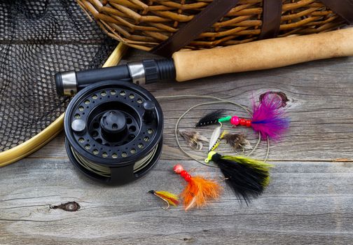 Close up top view of  fishing fly reel, landing net, creel and assorted flies, with partial cork handled pole on rustic wooden boards 