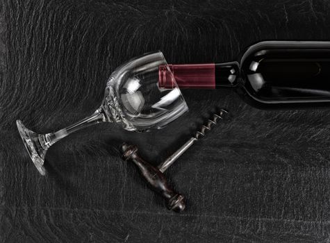 Overhead view of antique wine corkscrew, red wine bottle, and drinking glass on black slate 