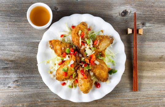 Top view of fried Asian style chicken wings in white plate with garnishes. Green tea and chopsticks in holder. Rustic wooden boards underneath. 