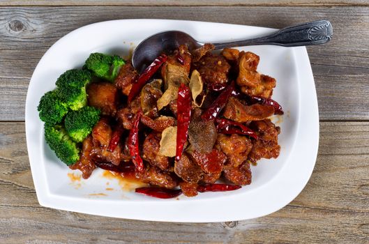 High angled view of Chinese dish consisting of fried tofu, red peppers, and broccoli. Large serving spoon on side of plate with rustic wood underneath. 