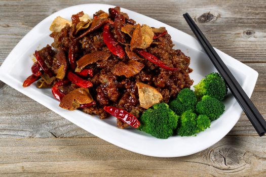 Close up front view of Chinese dish consisting of fried tofu, chicken, red peppers, and broccoli. Chopsticks on side of plate with rustic wood underneath. 