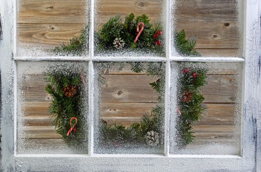 Snow covered window with decorative Christmas wreath on window with rustic wood in background. 