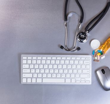 Overhead view of a medical stethoscope, computer keyboard, mouse and pills on stainless steel table