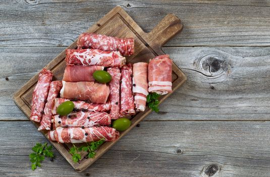 Top view horizontal image of various meats on serving board with ham, pork, beef, parsley, and olives on rustic wood. 
