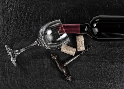 Overhead view of antique wine corkscrew, red wine bottle, glass and used corks on black slate 