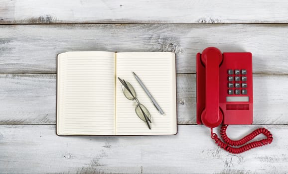 Vintage red phone, notepad, pen and reading glasses on rustic white wooden boards. High angled view. 

