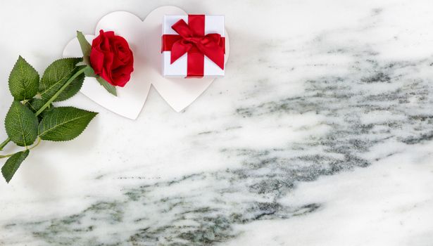 Single red rose resting on card and gift box on marble stone background in flat lay view 