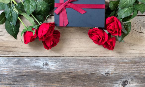 red roses surrounding dark gift box on weathered wooden background in flat lay view 