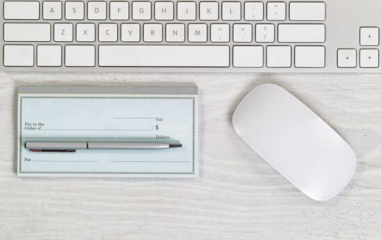 Image of partial keyboard, blank checkbook, silver pen and mouse on white desktop. Layout in horizontal format.