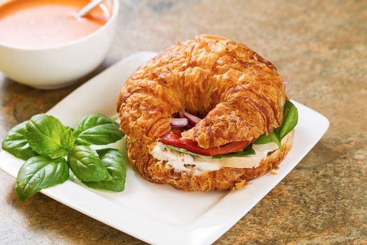 Horizontal photo of a chicken salad, tomato, basil, and onion inside croissant bread on white square plate with tomato soup and spoon in bowl all resting on natural stone countertop 