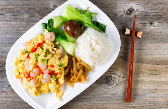 Top view of freshly cooked egg and shrimp dish with bok choy, soy sauce egg, cucumber, rice and chopsticks in holder on rustic wood. 