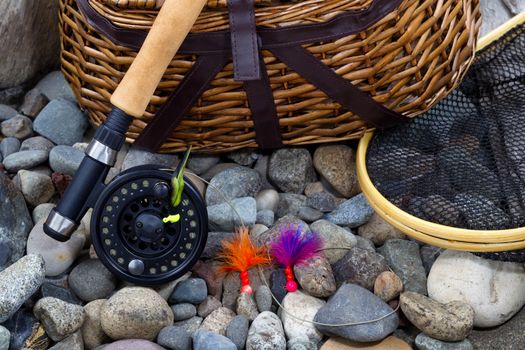 Top view up of  fishing fly reel, landing net, creel and assorted flies on dry river bed rocks 