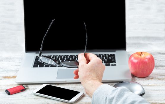 Close up of male hand holding reading glasses with workstation technology in background for business or education use 