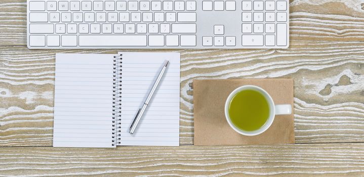 Top view shot of an old white desktop with keyboard, green tea in cup, notepad and pen in horizontal format.