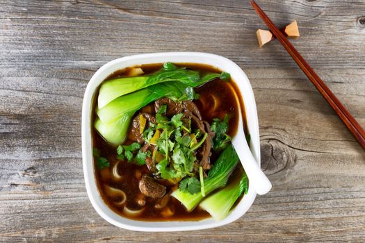 High angled view of beef noodle soup with bok choy and parsley. Soup in soup with chopsticks in holder on rustic wood.  