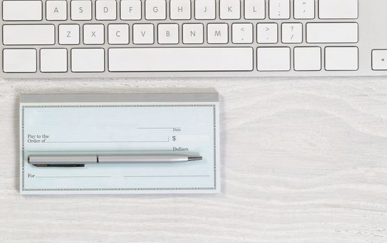 Image of partial keyboard with blank checkbook and silver pen on white desktop. Layout in horizontal format.