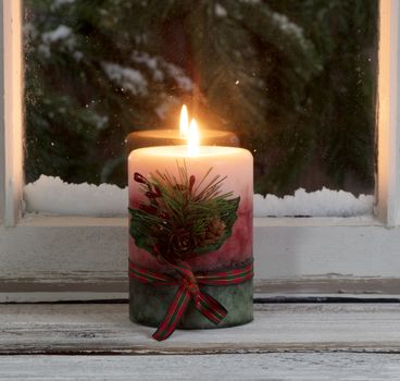 Christmas candle glowing on window sill with snowy evergreen branches in background 