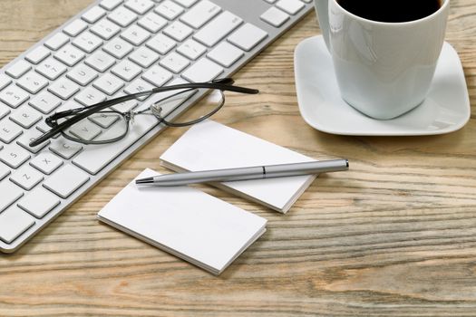 Close up of business cards and silver pen. Computer keyboard, reading glasses and cup of coffee in background on desktop. 