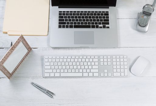 Top view angle of desktop layout for proper use consisting of laptop, keyboard, pens, mouse, picture frame, phone, and work folders on white desk.